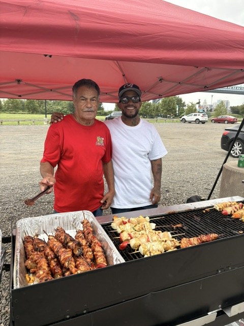 Two CoquiEats cooks grilling trays of pinchos under a red canopy